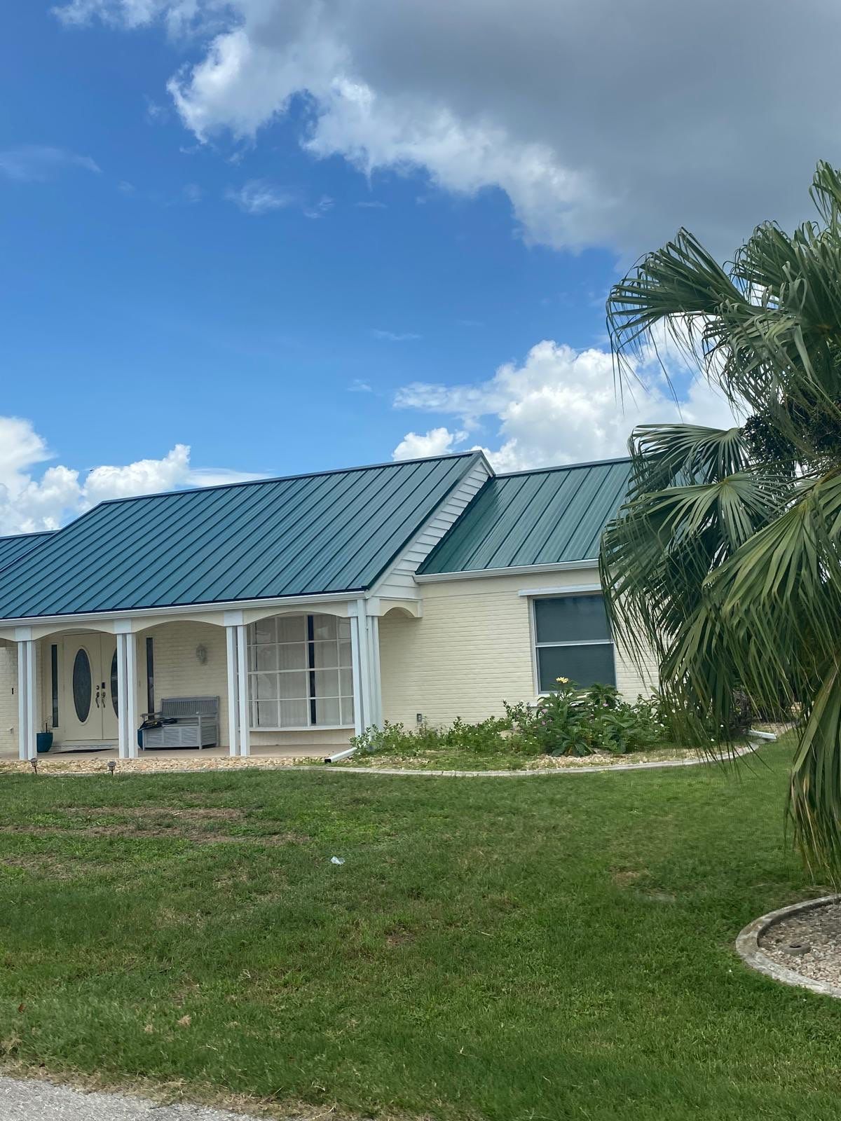 A cream-colored house with a teal metal roof under a blue sky, palm tree on right.