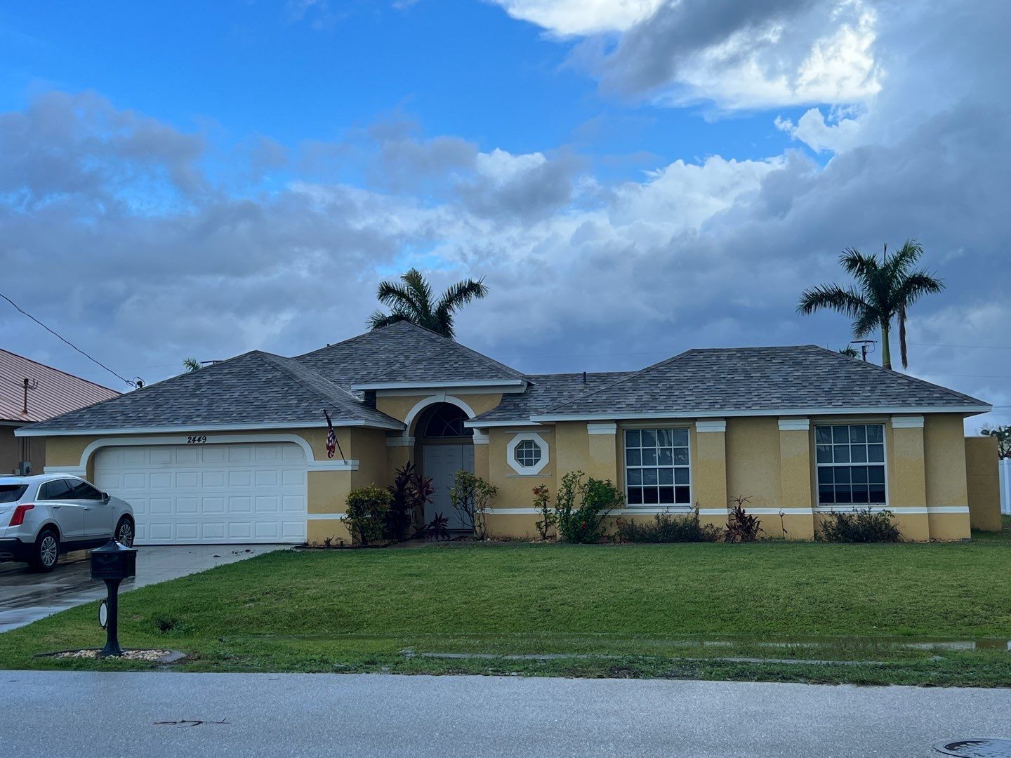 Yellow house with a white garage door, palm trees, and a car in the driveway under a cloudy sky.