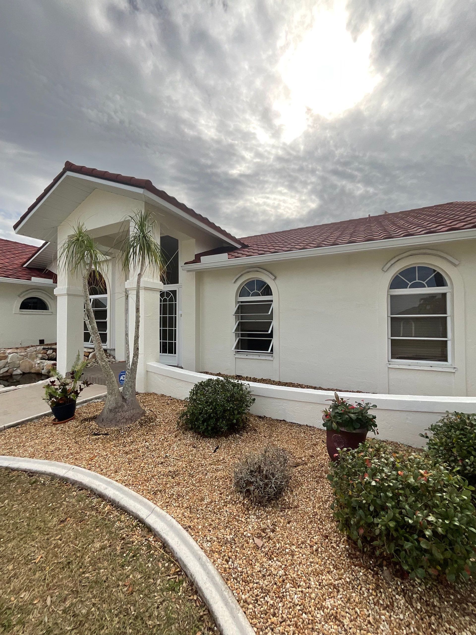 White stucco house with red tile roof, arched windows, and landscaping under cloudy sky.
