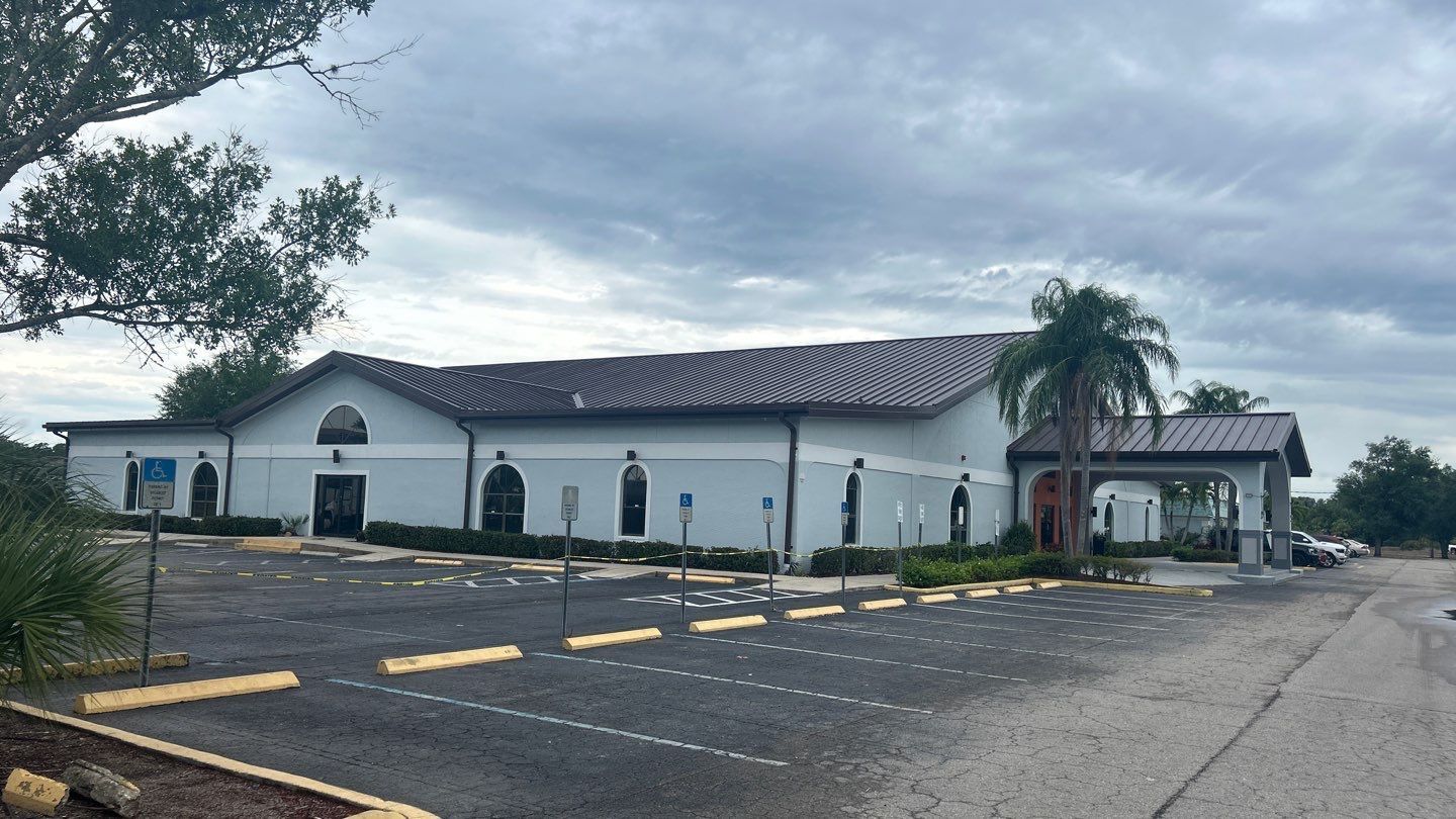 Exterior view of a light blue building with a dark brown roof and a paved parking lot. Cloudy sky.
