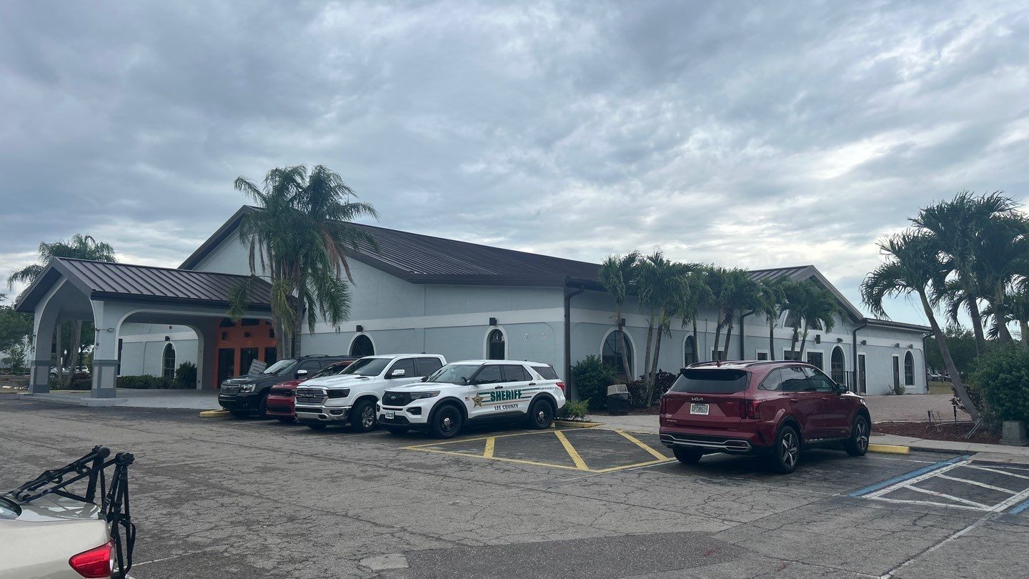 Exterior view of a building with a line of parked police cars. Cloudy sky, palm trees.
