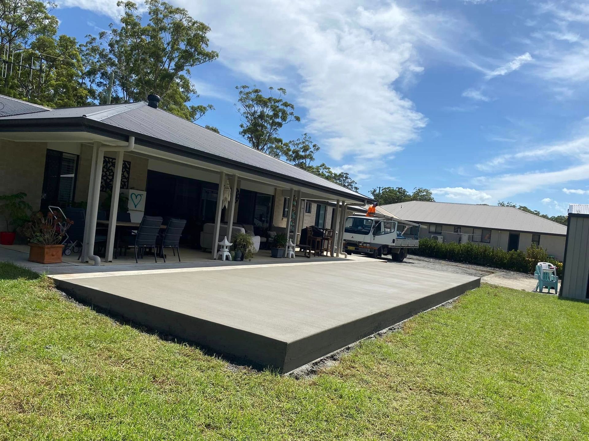 Newly poured concrete patio next to a house with a grey roof and lush green grass on a sunny day — Rhyno’s Concreting In Old Bar, NSW