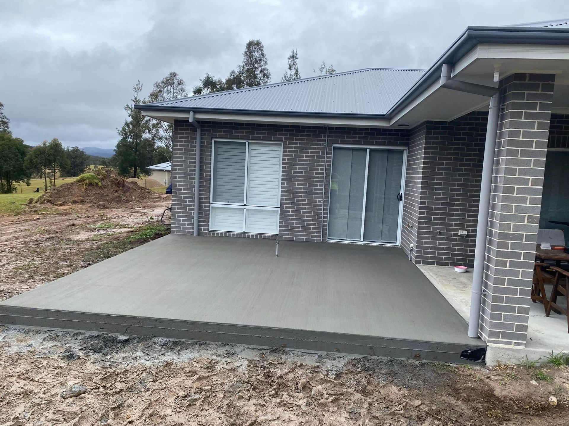 A Freshly Poured Concrete Patio Next to A Brick House with Sliding Glass Doors Under an Overcast Sky — Rhyno’s Concreting In Old Bar, NSW