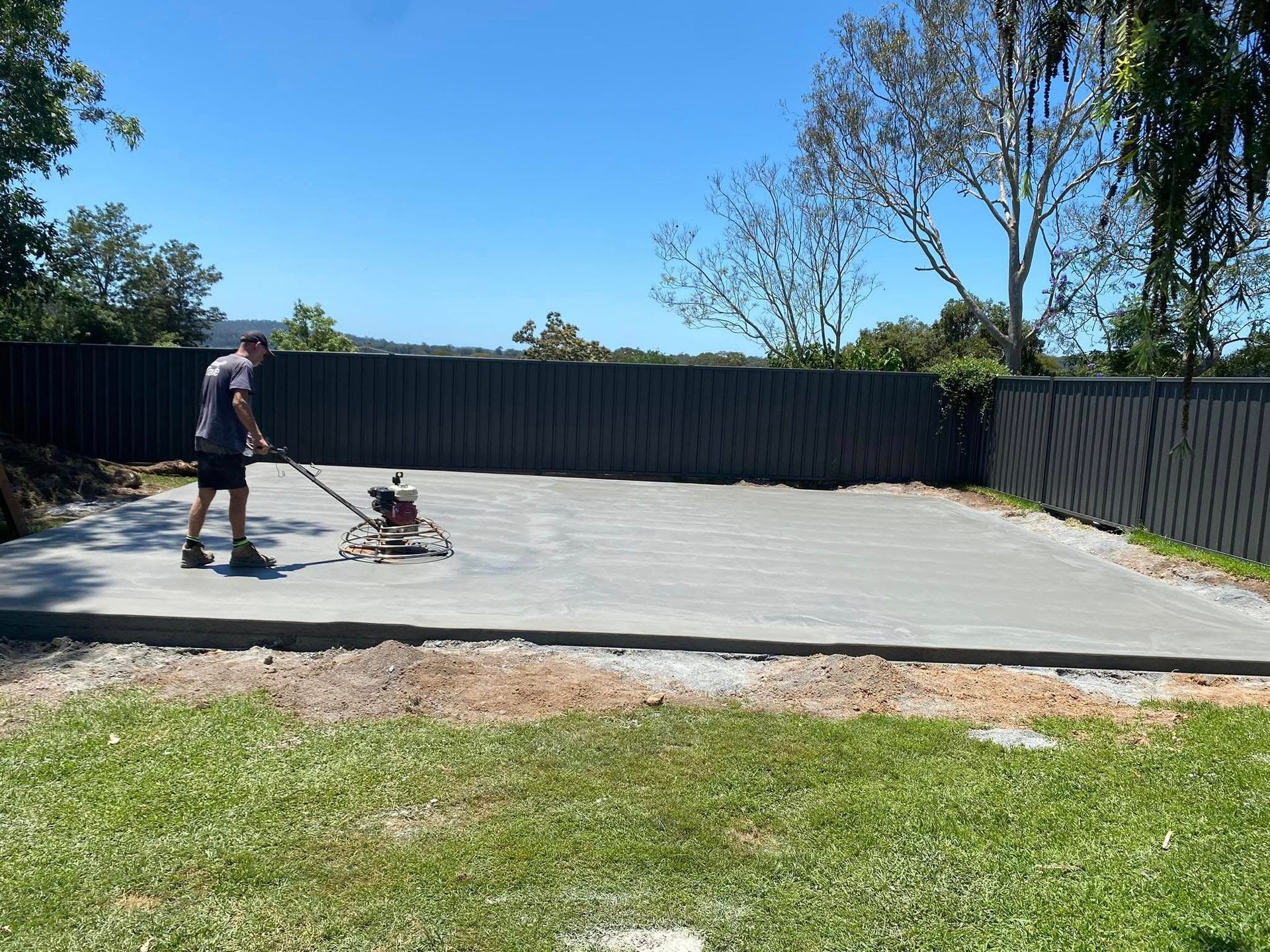 Man using a power trowel on a newly poured concrete slab in a backyard on a sunny day — Rhyno’s Concreting In Old Bar, NSW
