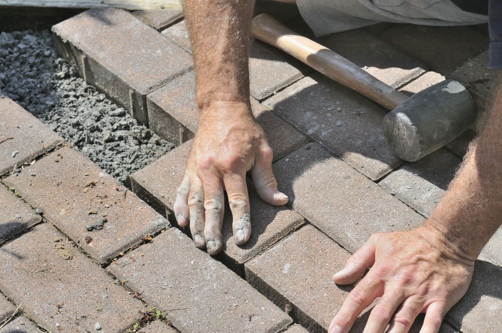 Hands laying brick pavers with a hammer in the background — Rhyno’s Concreting In Old Bar, NSW