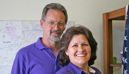 Two movers in blue coveralls lift a bubble-wrapped box in a room with furniture and boxes