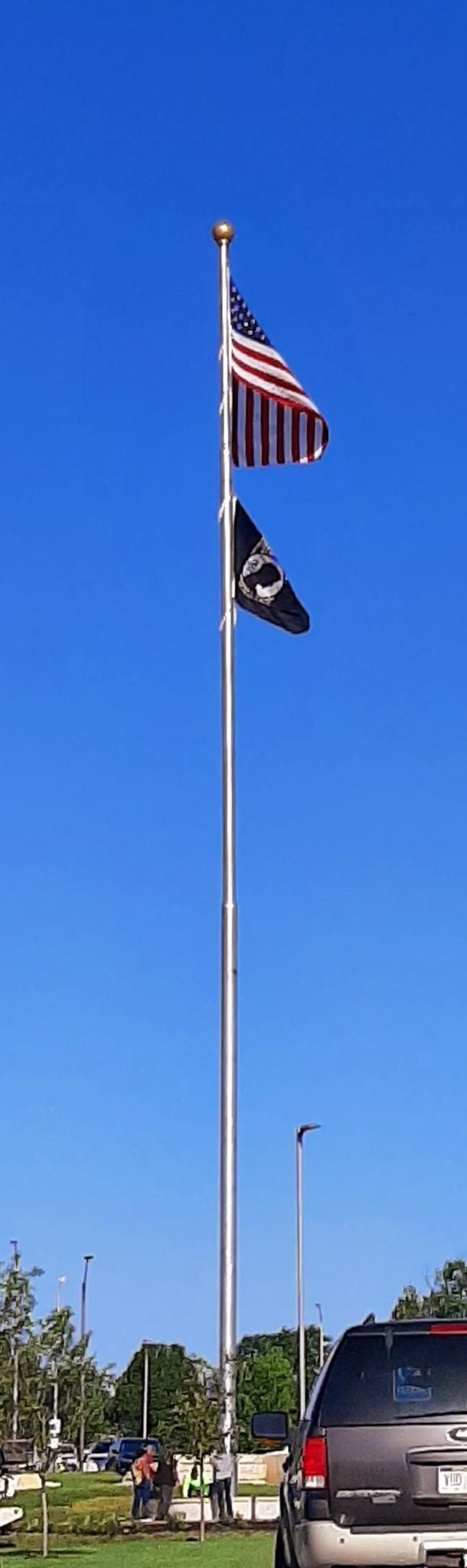 A car is parked in front of a tall flag pole with two flags on it.