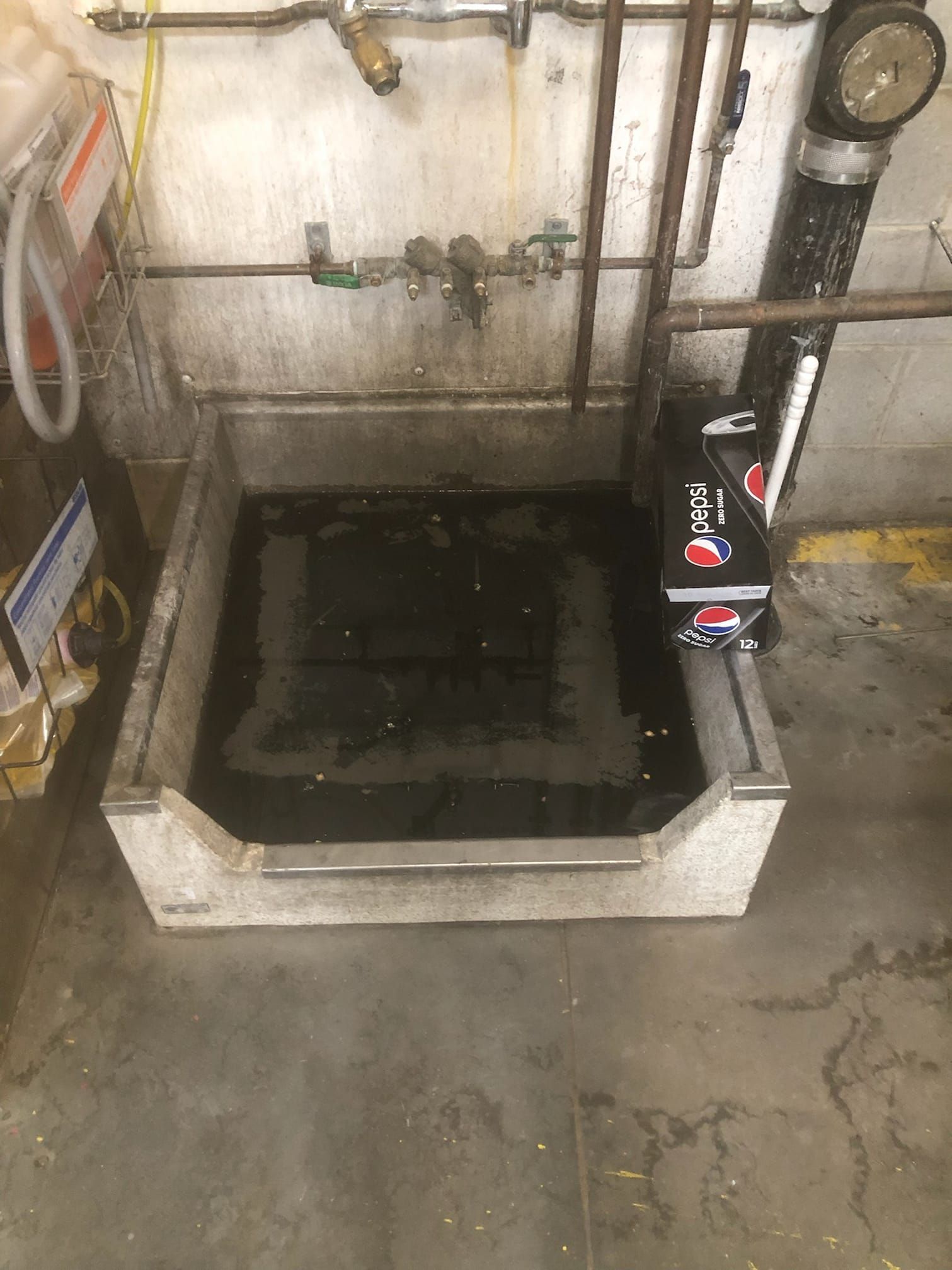 A utility sink filled with dark water, located in a commercial setting, with pipes and boxes nearby.