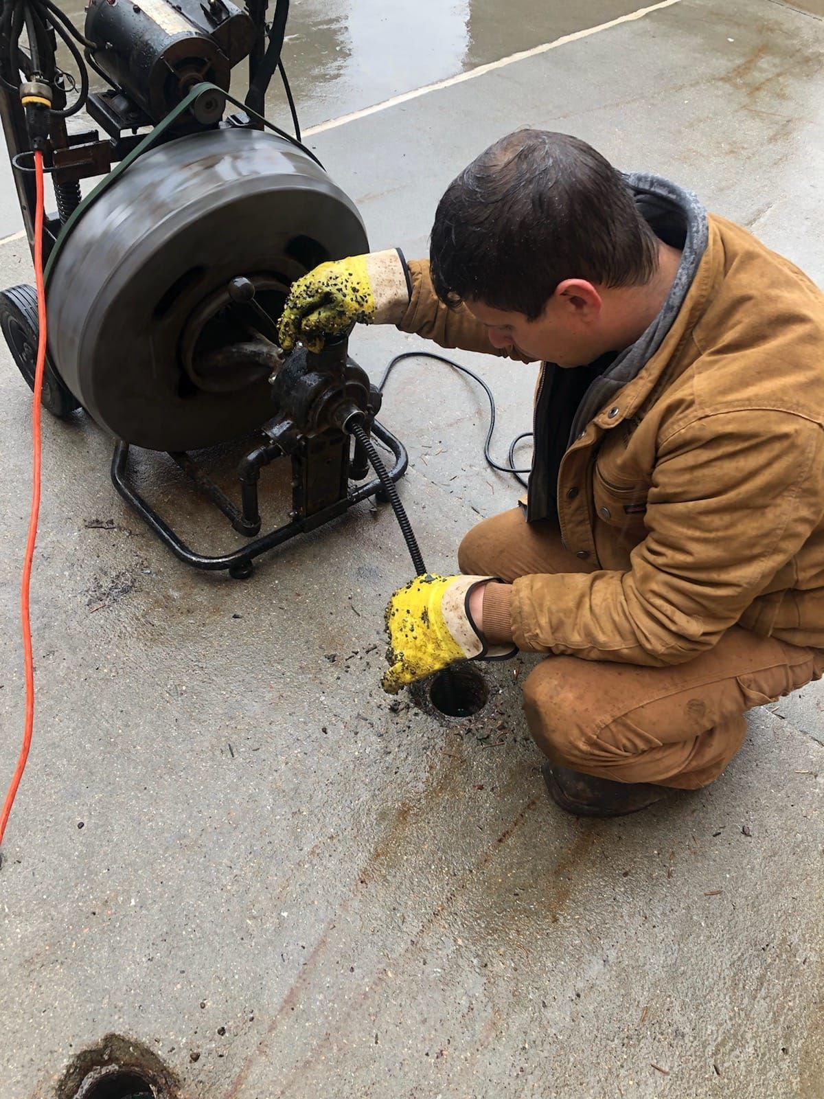A plumber in a brown jacket uses a drain cleaning machine outside on concrete.