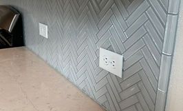 Herringbone-patterned gray tile backsplash with two electrical outlets, installed above a tan countertop.