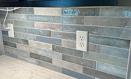 Blue and gray rectangular tile backsplash with two white electrical outlets.