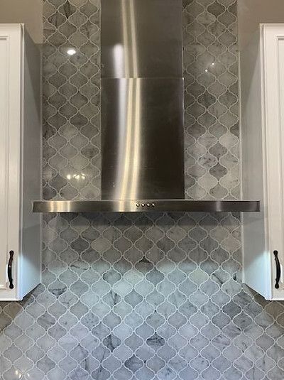Stainless steel range hood centered on gray patterned tiled backsplash. White cabinets on each side.