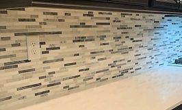 Close-up of a kitchen backsplash with horizontal mosaic tiles in white, gray, and brown. White countertop below.