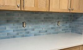 Blue tiled kitchen backsplash with two electrical outlets, beneath wood cabinets and countertop.