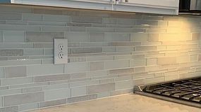 Off-white rectangular tiles on kitchen backsplash with a white outlet, stove, and cabinet.