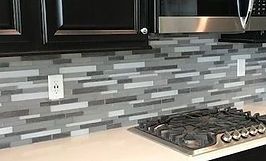 Kitchen backsplash with gray, white, and dark gray rectangular tiles, below black cabinets and a microwave.