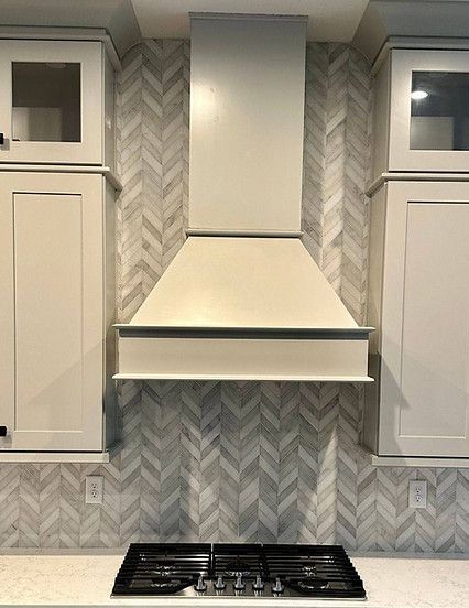 White kitchen hood with cabinets and herringbone tile backsplash over a stovetop.