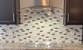 Kitchen backsplash with a pattern of gray and white rectangular tiles, beneath a stainless steel hood and dark cabinets.