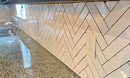 Herringbone pattern white tiled kitchen backsplash, with granite countertop and electrical outlet.