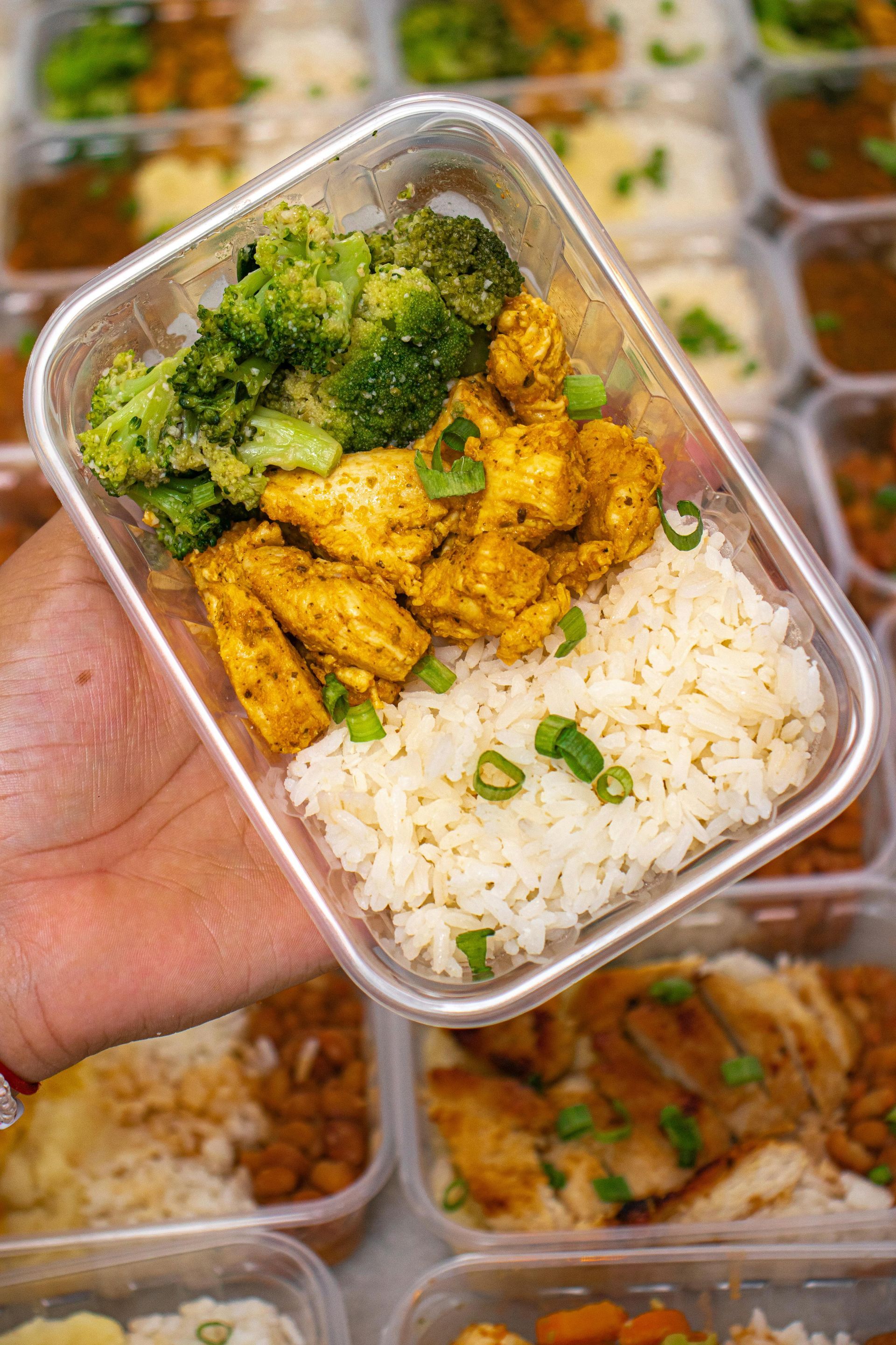 A hand holds a plastic meal prep container filled with yellow seasoned chicken, broccoli, and white rice with green onion.