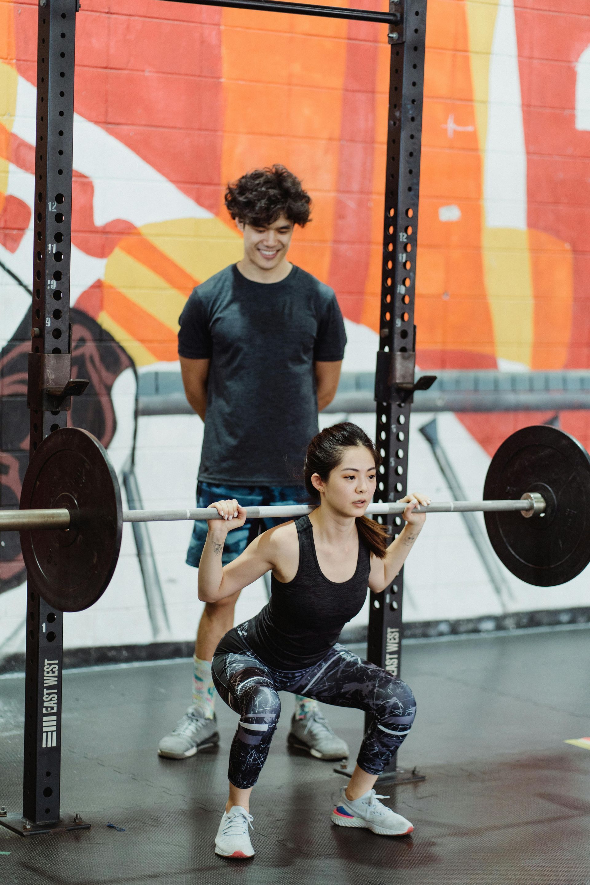 Woman doing a barbell squat while a coach spots her in a gym with an orange mural background