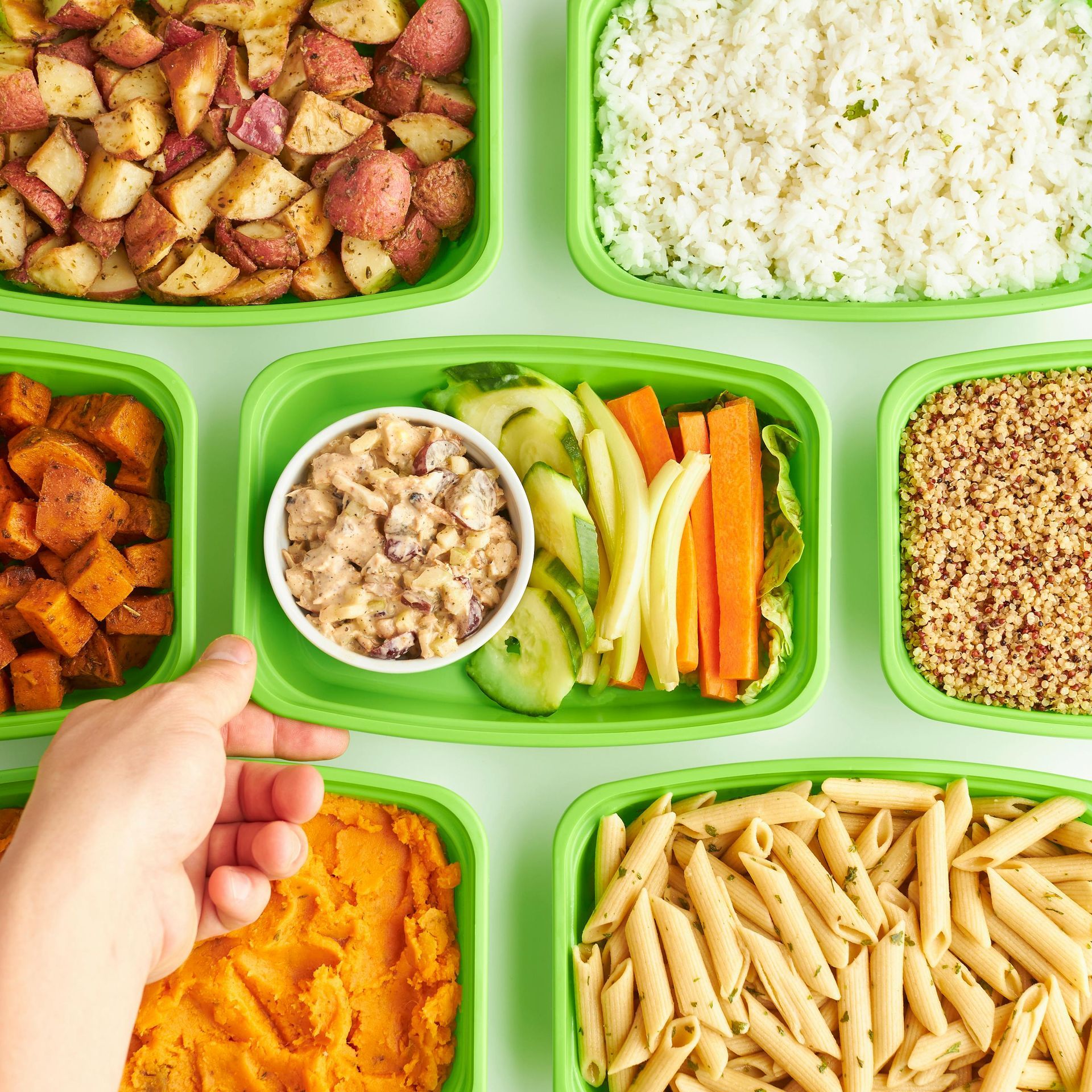 Hand placing a green container of dip and veggies among various other meal prep boxes filled with grains, pasta, and sides.