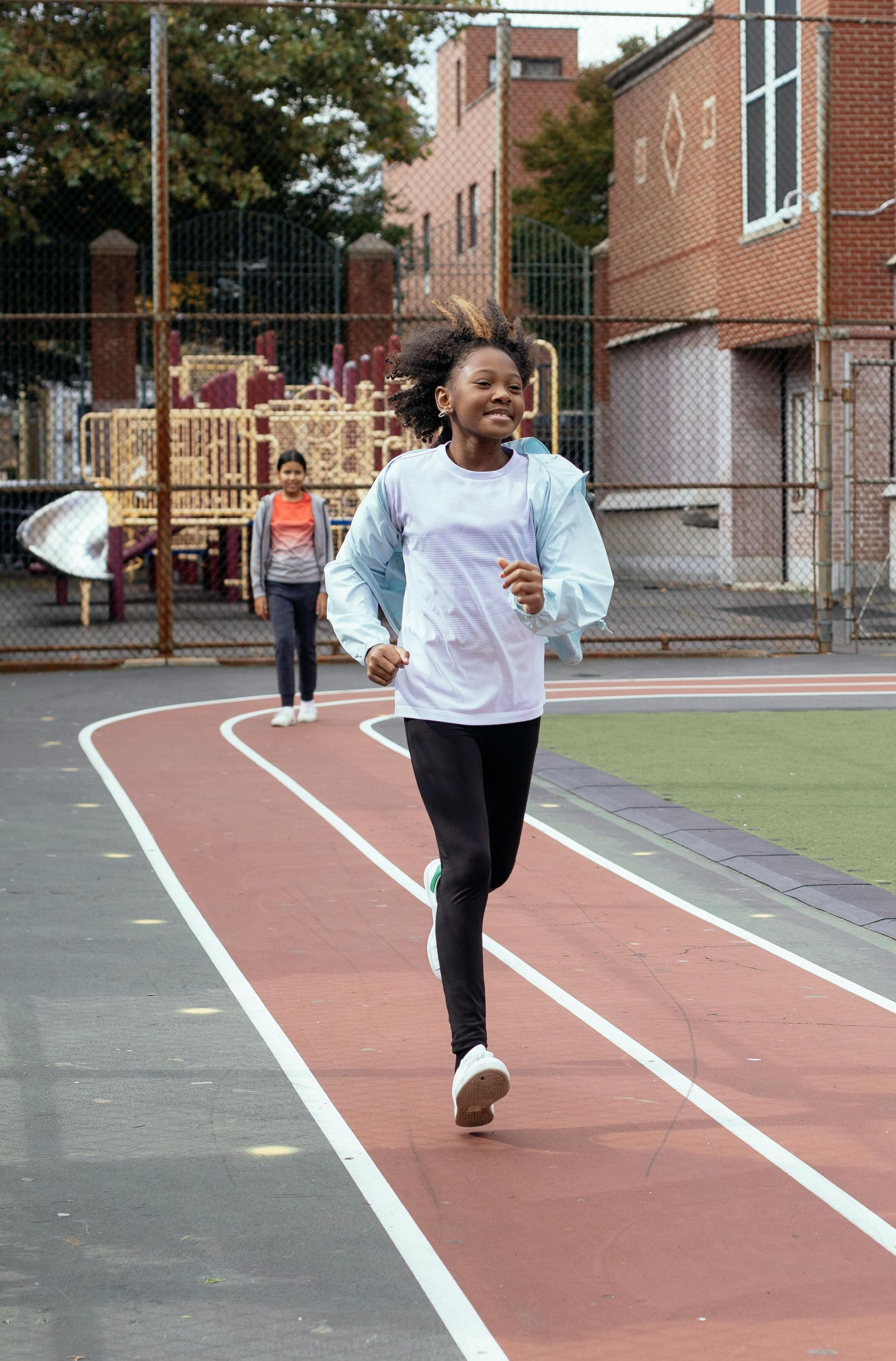 Runner in white shirt jogging on an outdoor track, with another person behind near school buildings.
