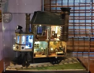 Miniature lit house model with warm interior lights and a blue-gray exterior on a wooden base near blinds.