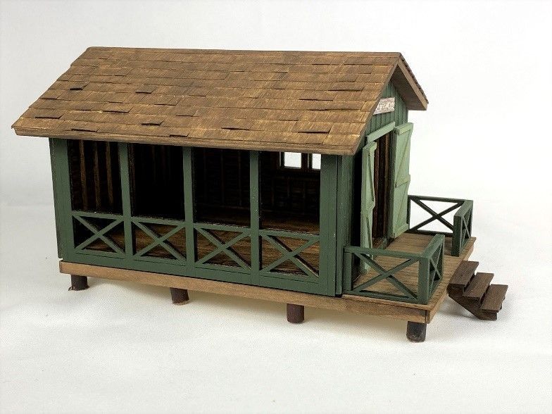 Small green wooden model train station with a covered porch and shingled roof