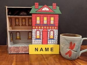 Miniature colorful house with a yellow nameplate and a white mug with red flowers on a table