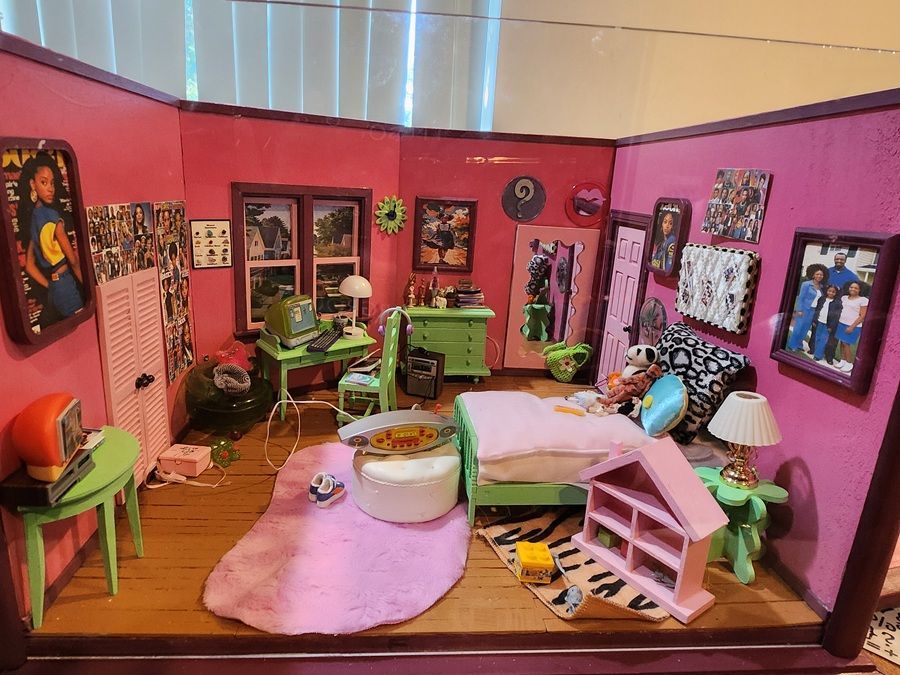 Pink dollhouse room with green furniture, wall art, and a white bed on a pink rug