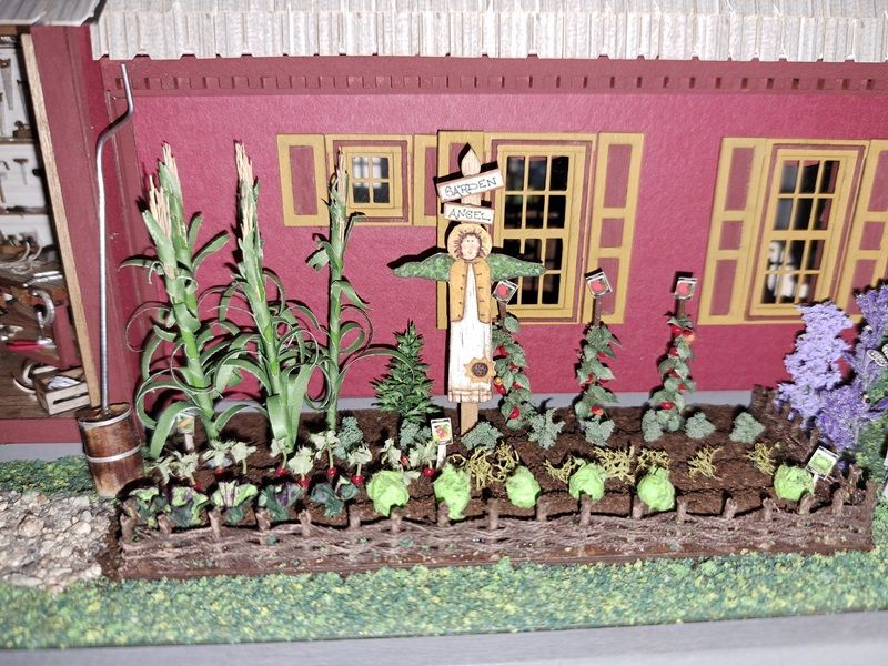 Miniature garden beside a pink house wall with potted flowers, greenery, and a small scarecrow decoration