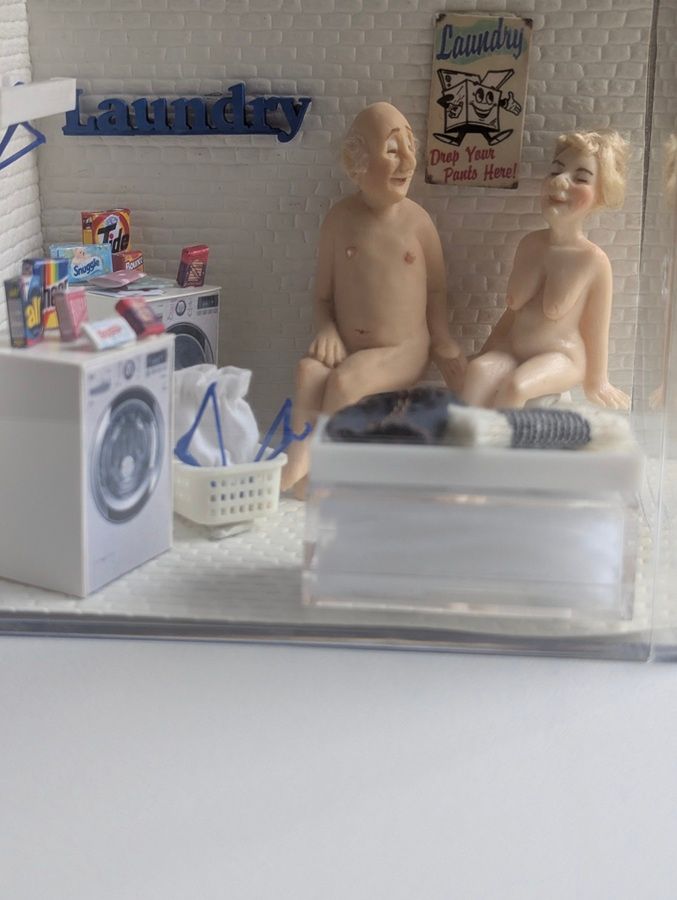 Miniature laundry room with washer, basket, and two nude doll figures seated on the floor