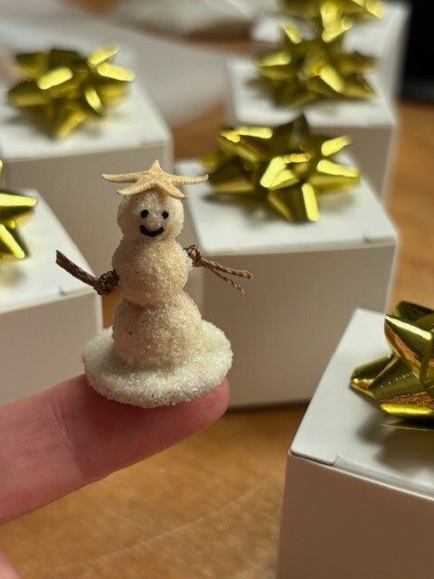 Tiny snowman figurine on a fingertip, with gold gift boxes in the background.
