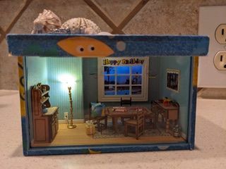 Miniature dollhouse room with blue walls, a window, table, chairs, lamp, and small cabinet.