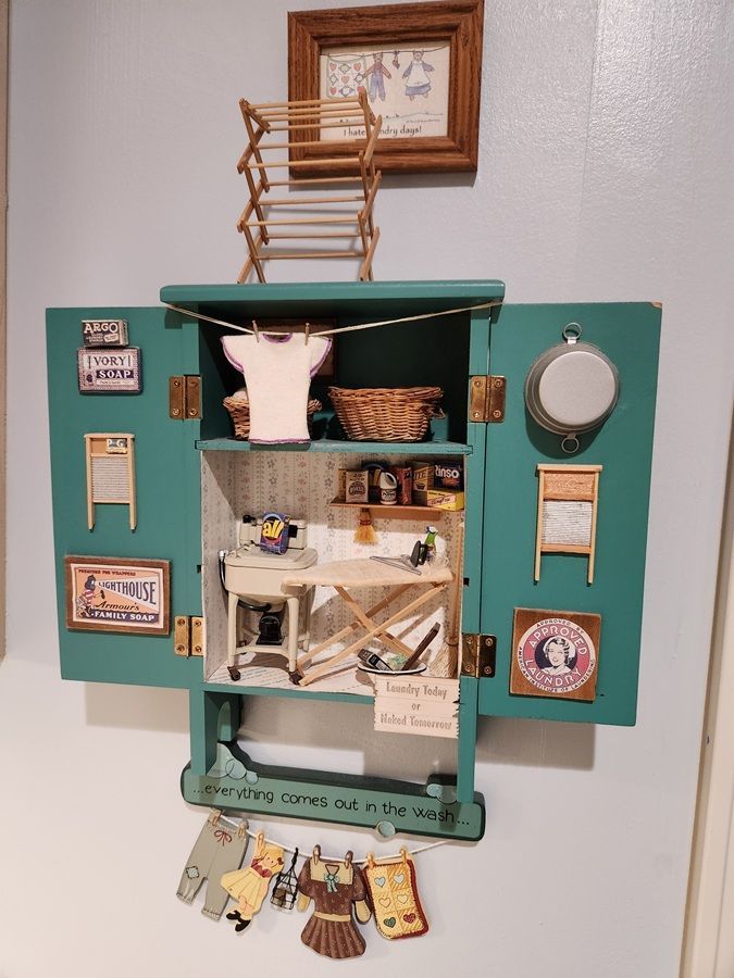Teal wall-mounted miniature shop display with tiny shelves, framed art, and hanging decorations