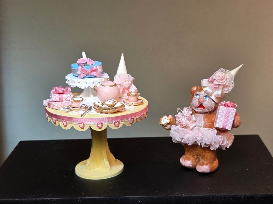 Two pastel doll figurines on a table, one on a cake stand and one in a pink dress with gifts.