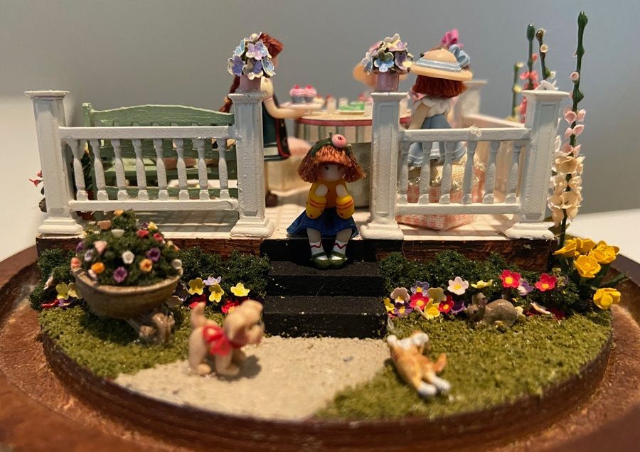 Miniature garden scene with white fence, flowers, small porch, and tiny animals on a grassy oval base.