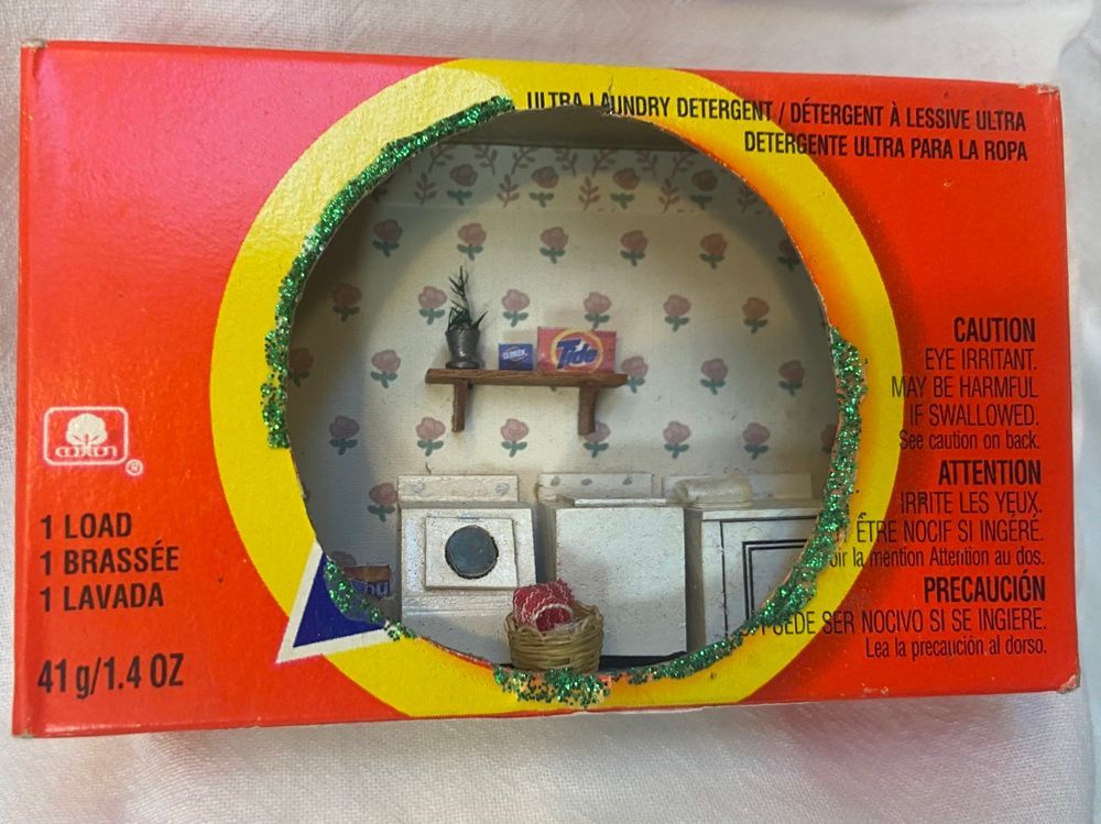 Orange washing powder box with a round window showing a laundry room scene