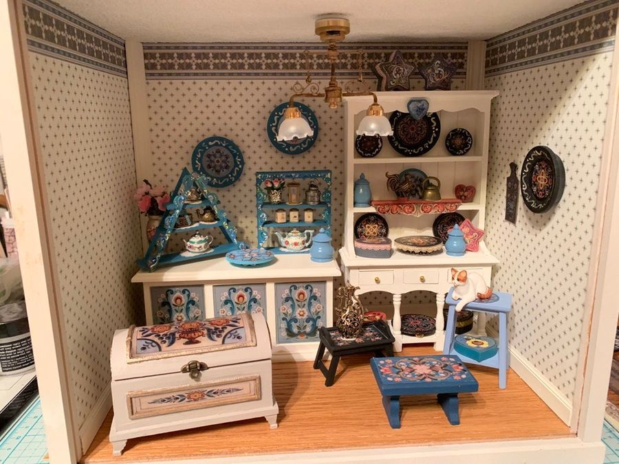 Miniature dollhouse room with blue-and-white furniture, shelves of dishes, and patterned wallpaper