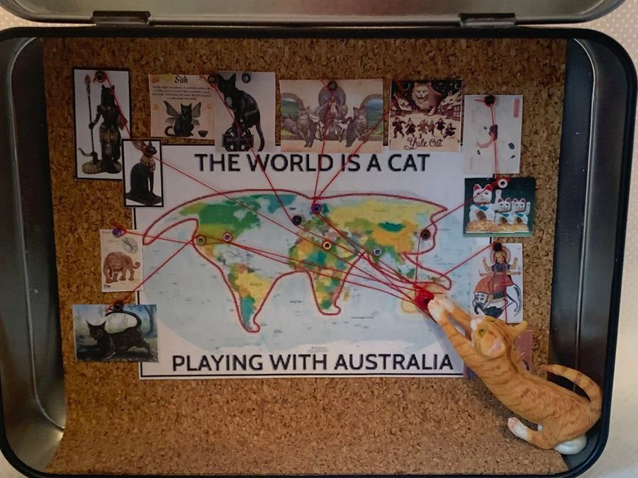 Corkboard display titled “The World Is a Cat, Playing with Australia” with maps, photos, and string lines