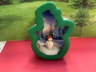 Green tree-shaped display with a small snowman figurine inside on a red surface outdoors