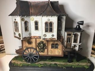 Miniature rustic house model with white and tan walls, dark roof, and a large wagon wheel on a grassy base