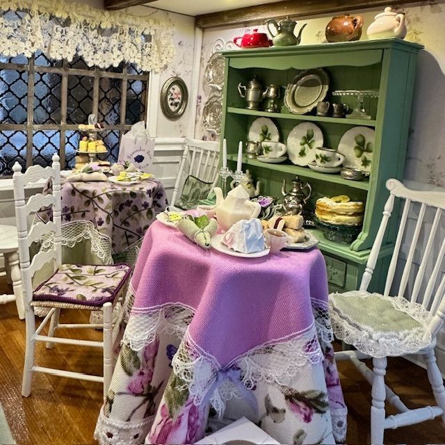 Cozy vintage dining nook with lace-covered table, white chairs, floral china cabinet, and floral decor.