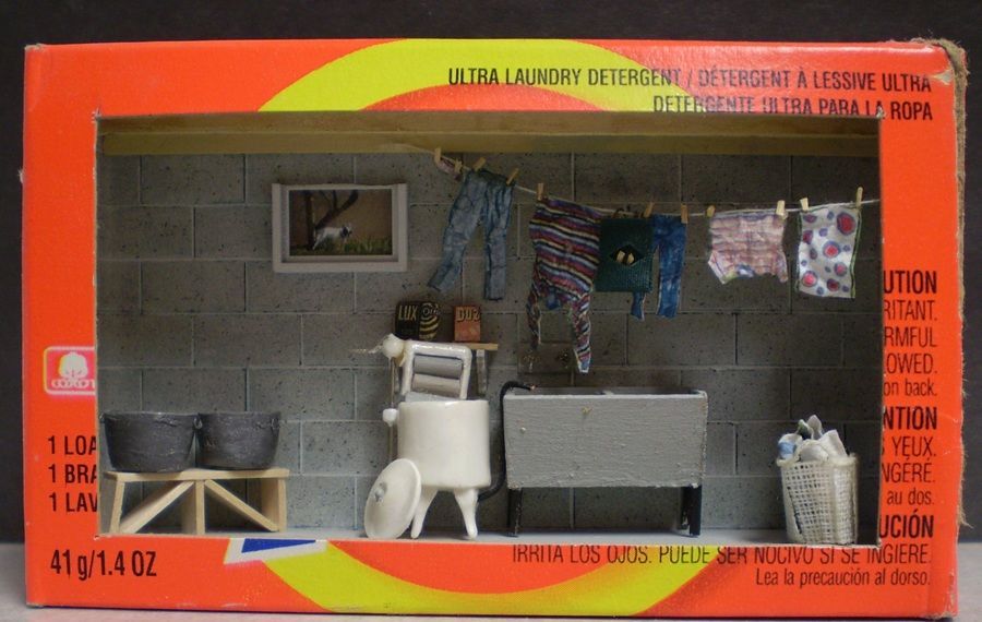 Miniature laundry room scene in a boxed set with washer, sink, hanging clothes, and cleaning items.