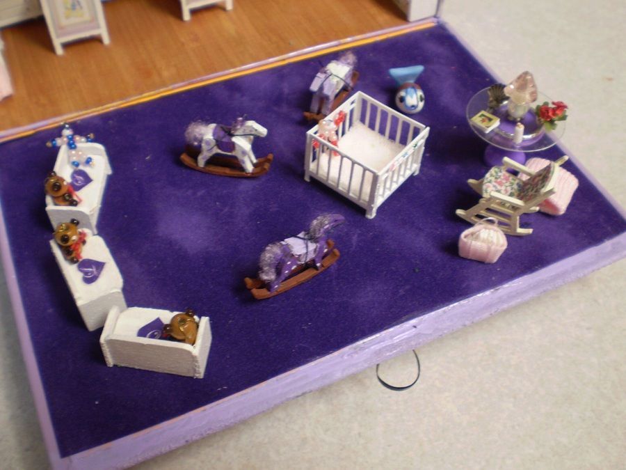 Miniature toy room display on a purple base with small figures, furniture, and a crib-like bed