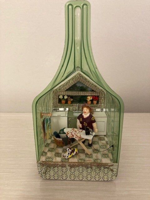 Miniature dollhouse scene in a green bottle-shaped frame, with a seated figure in a cozy room.