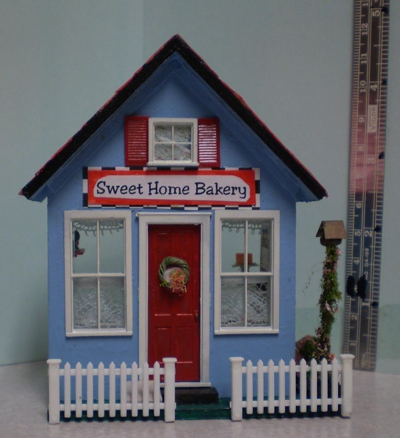 Miniature blue bakery storefront with red door, white picket fence, and “Sweet Home Bakery” sign