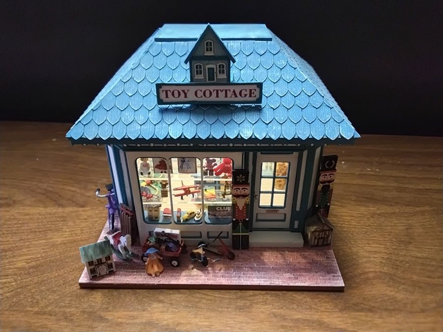 Miniature toy cottage with blue roof, lit windows, and tiny figures on a wooden base.