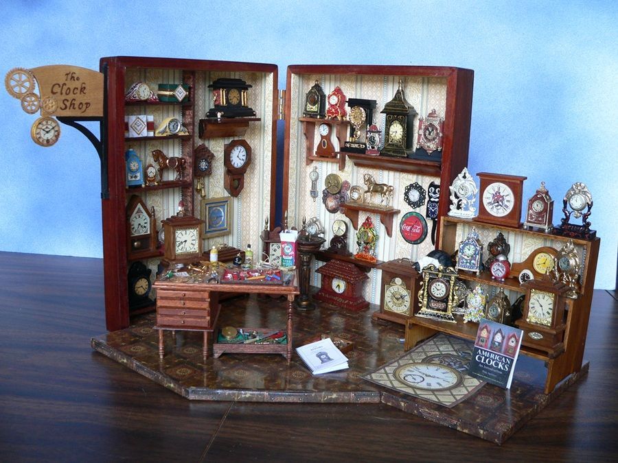 Miniature dollhouse room with shelves, clocks, framed art, and a desk in a rustic display.